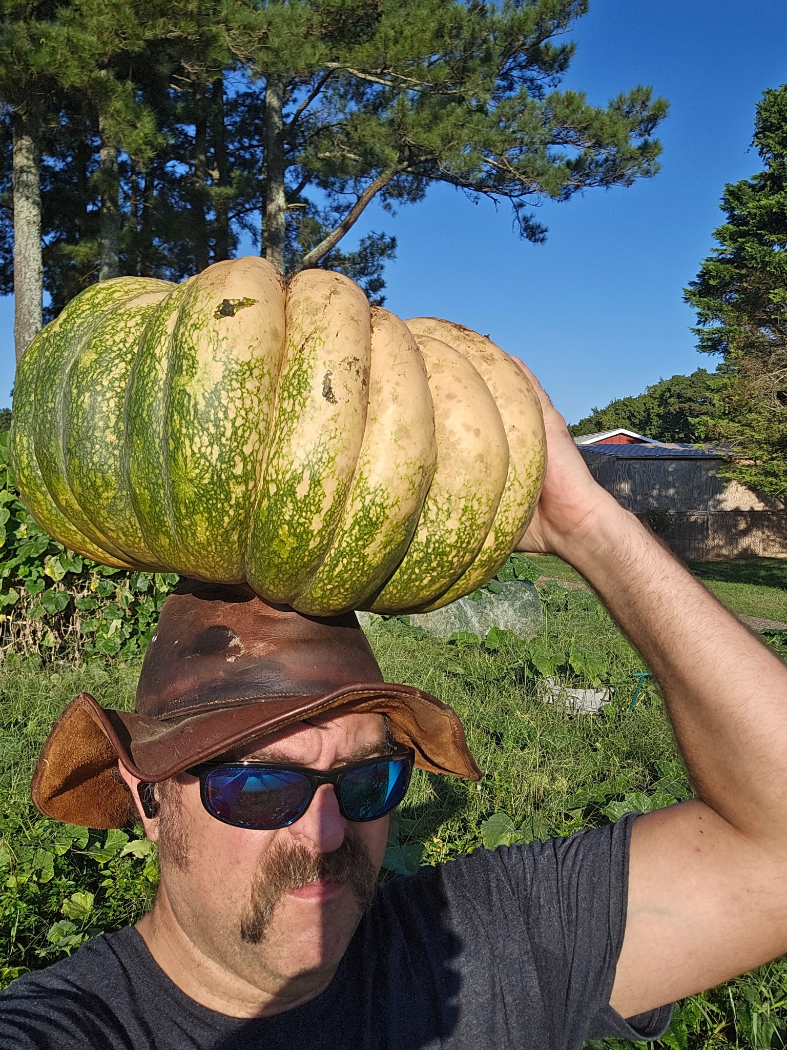 The owner of Creasy Aviation Farms holding a large green-and-tan pumpkin above his hat in the garden.